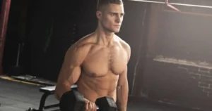 A shirtless, muscular man performing a dumbbell workout in a dimly lit gym