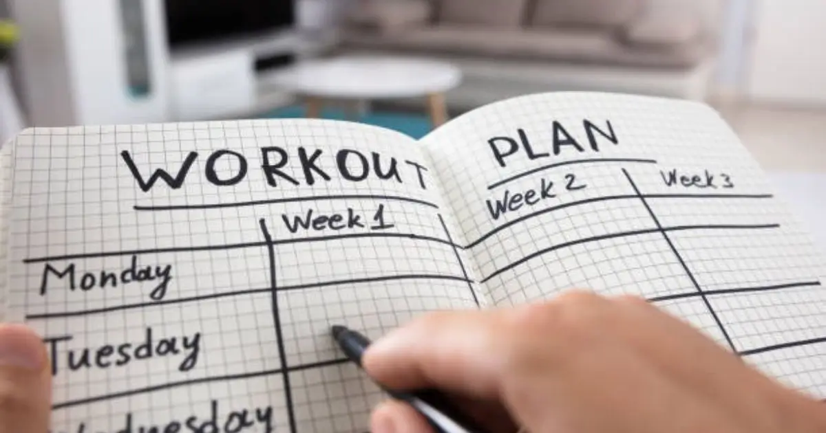 A person writing in a workout plan journal with a black pen, organizing fitness goals for multiple weeks.
