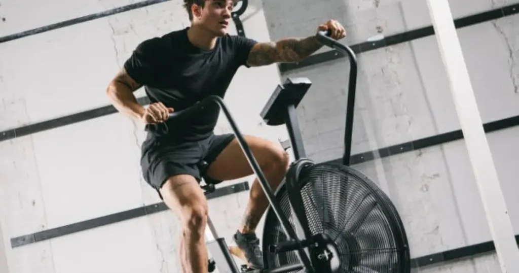 A fit man performing an elliptical HIIT workout on an air bike in a modern gym with an industrial-style design.