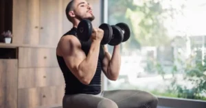 A fit man in a black sleeveless shirt performing a seated dumbbell workout at home.