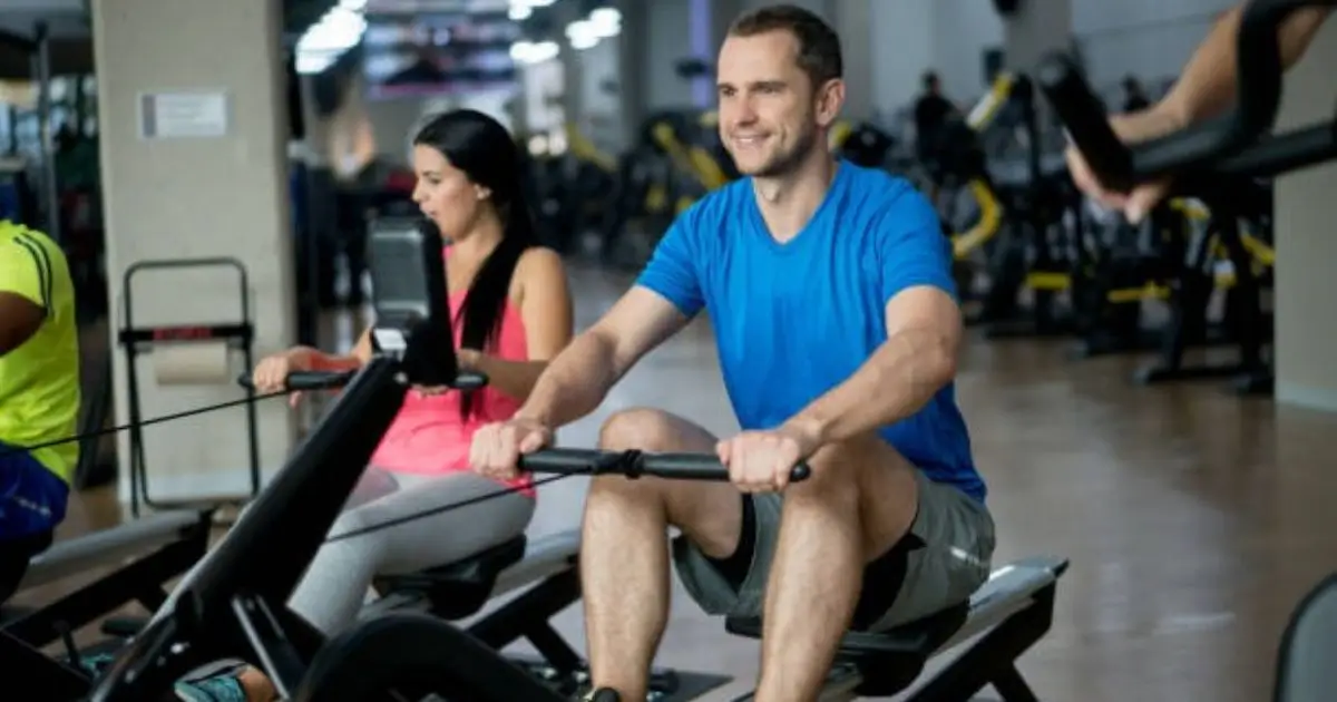 A smiling man in a blue shirt using a rowing machine at the gym, surrounded by other people working out.