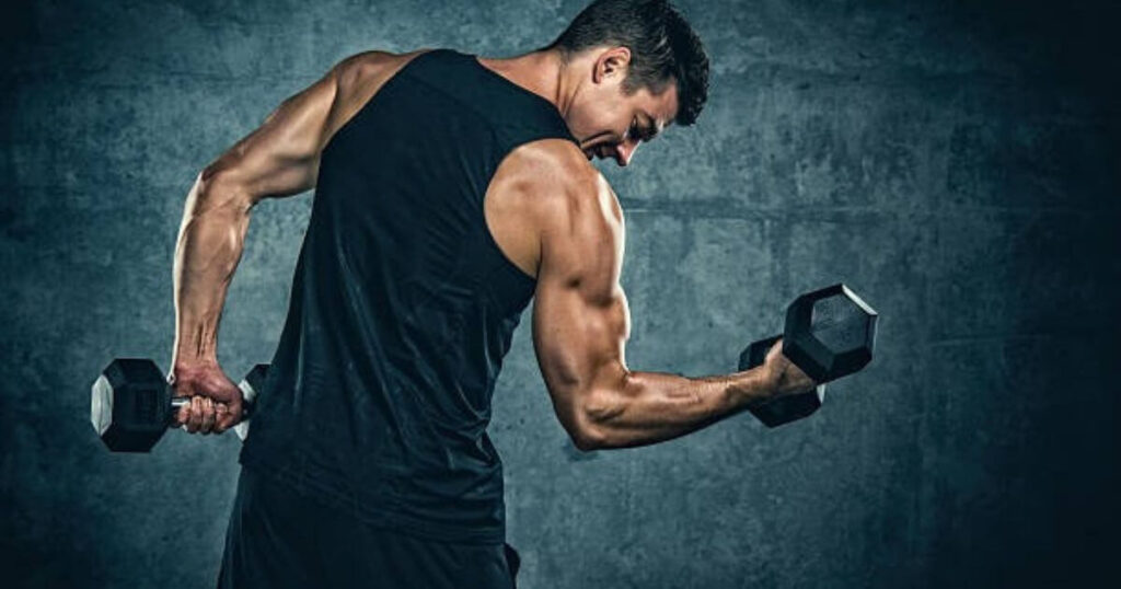 A muscular man in a black tank top performing a bicep curl with dumbbells, showcasing his toned arms and strong physique.
