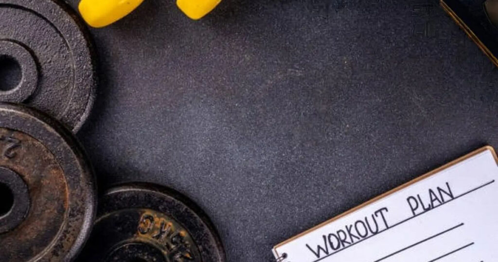 A workout plan notebook alongside black weight plates and yellow dumbbells on a dark surface.
