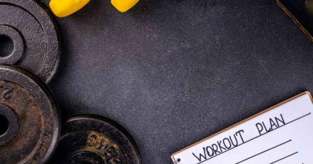 A workout plan notebook alongside black weight plates and yellow dumbbells on a dark surface.