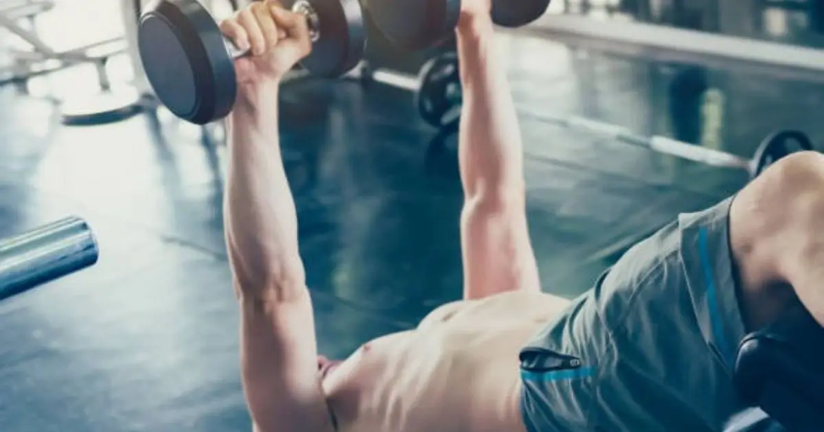 A shirtless man performing a dumbbell bench press in a gym.