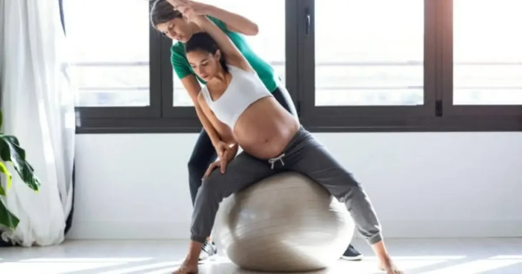 A pregnant woman exercising with a physiotherapist using a stability ball in a bright room.