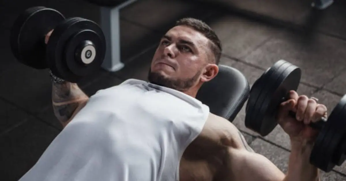 A muscular man in a white tank top performing an incline dumbbell press on a bench in a gym.