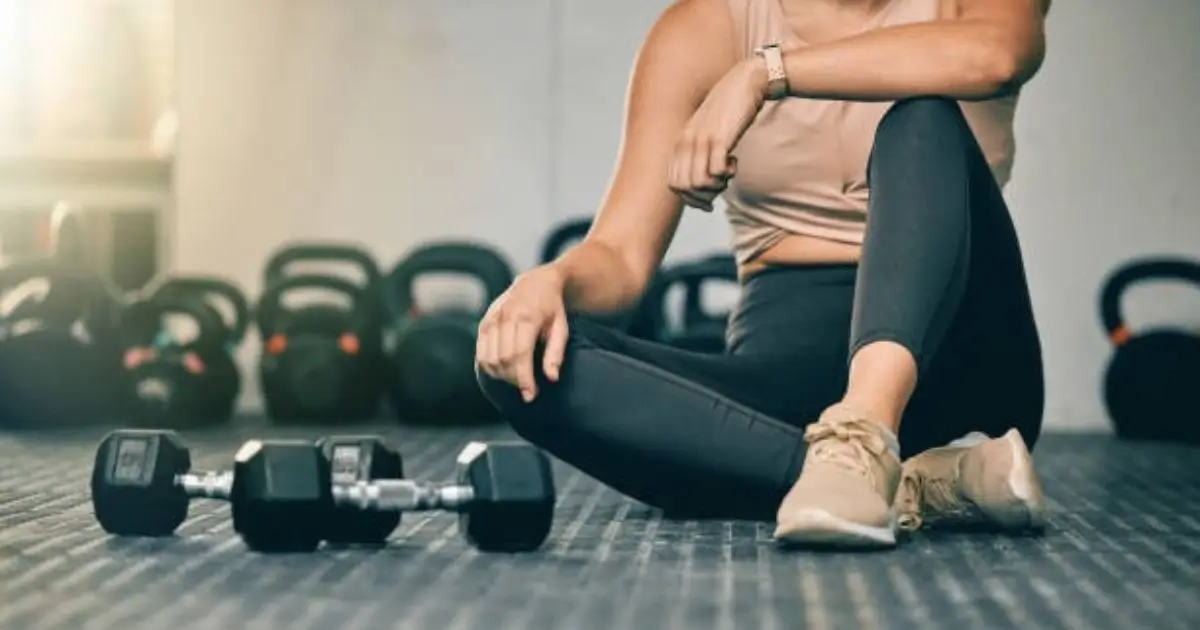 A woman in athletic wear sitting on the gym floor with dumbbells in front of her, surrounded by kettlebells in the background.