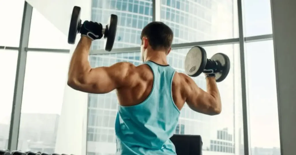 an in a blue tank top performing a dumbbell shoulder press in a gym with large windows overlooking a cityscape.