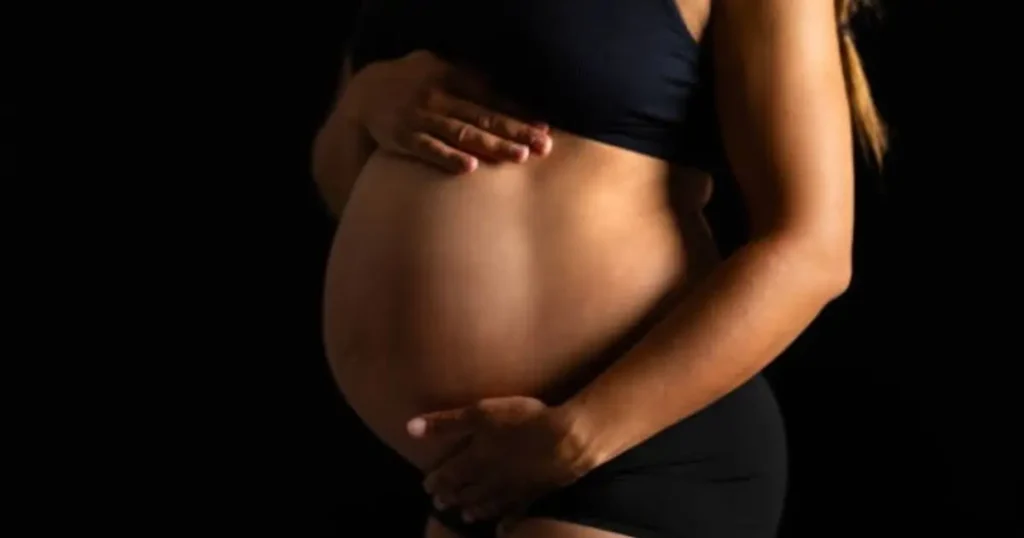A pregnant woman in black sportswear gently cradling her belly against a dark background.