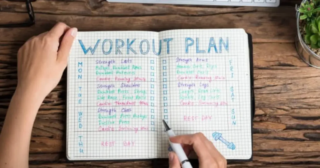 A handwritten workout plan in a notebook with different exercises listed for each day of the week, placed on a wooden desk with a pen, laptop, and a small plant nearby.