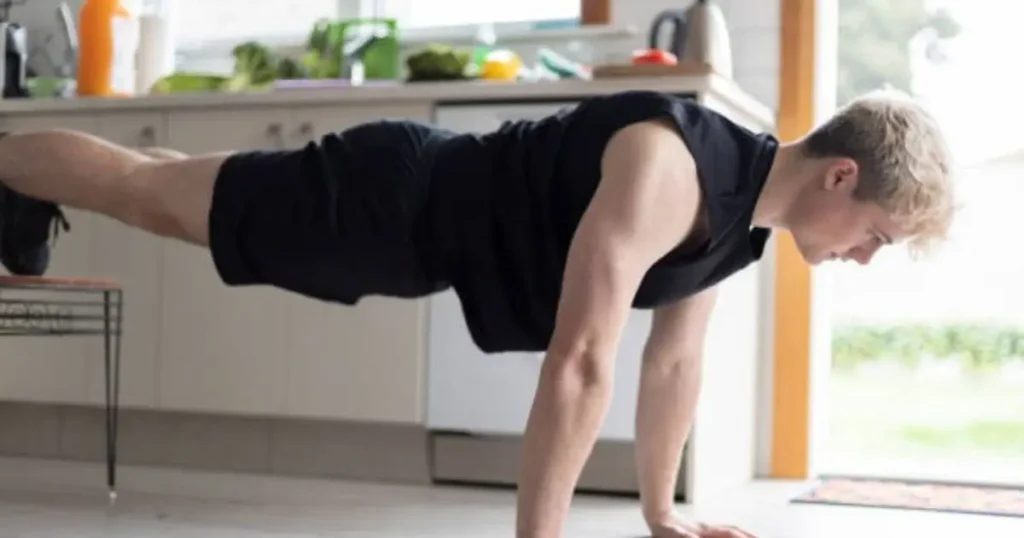 A young man performing a calisthenics workout in a kitchen, balancing in a plank position with his feet elevated.