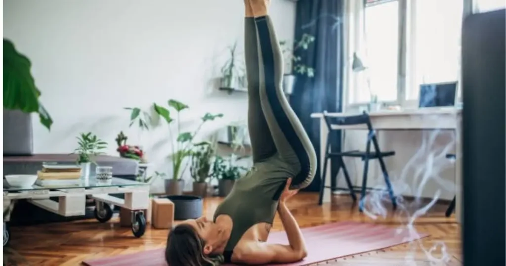 A woman practicing a lower back workout at home, performing a shoulder stand yoga pose on a pink mat in a cozy living room with plants and natural light.