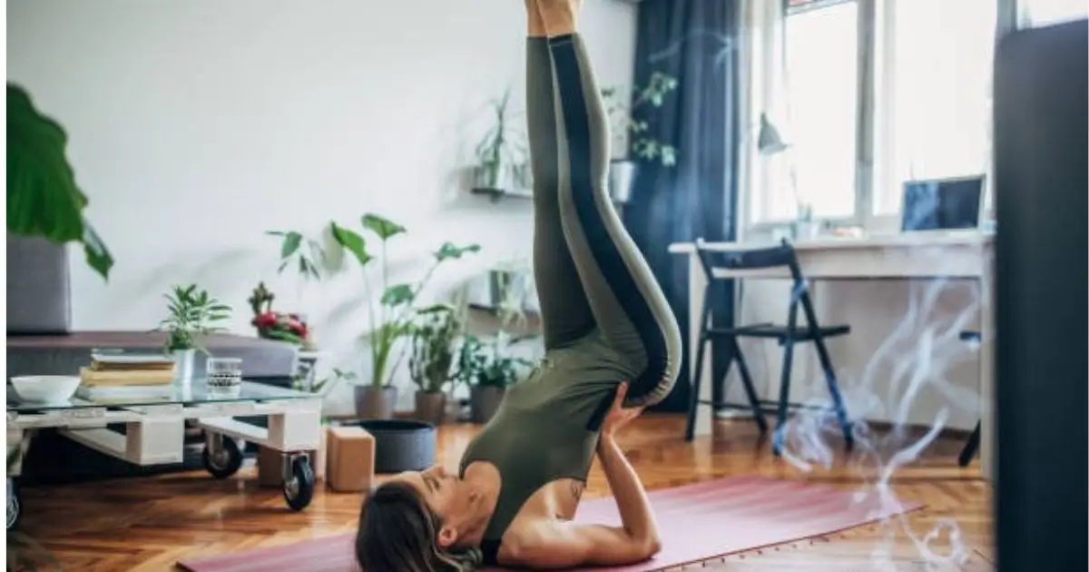 A woman practicing a lower back workout at home, performing a shoulder stand yoga pose on a pink mat in a cozy living room with plants and natural light.
