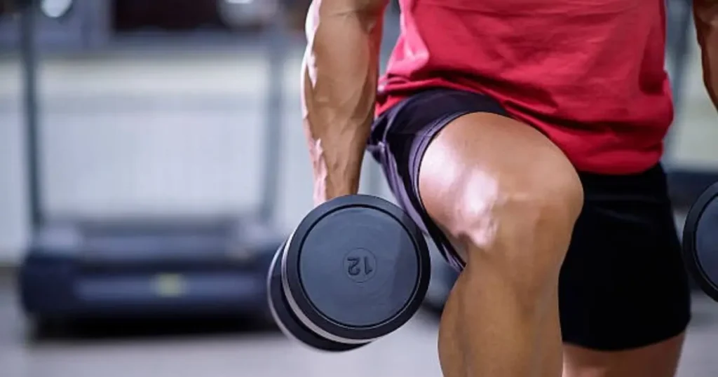 A person in a red shirt and black shorts is performing a seated dumbbell exercise, focusing on their legs and core.