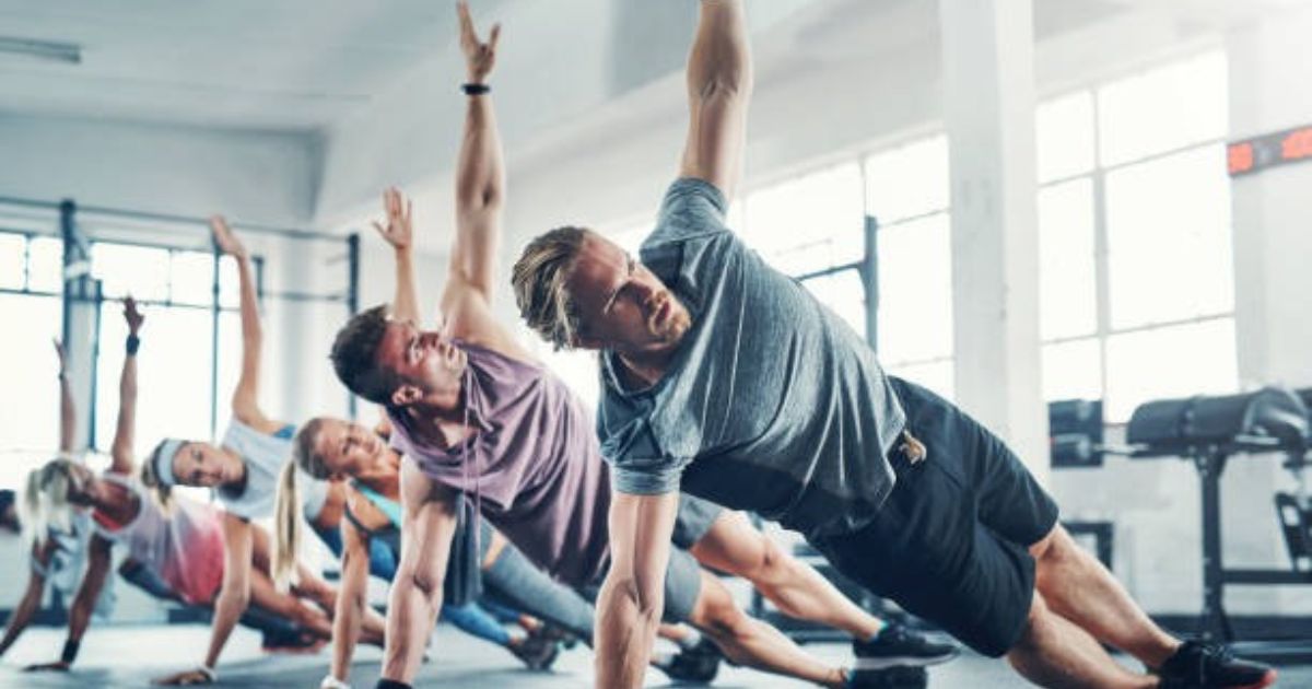 A group of people in a gym are performing synchronized side plank exercises, with one person leading the movement. The setting is bright and spacious, featuring large windows and exercise equipment in the background.