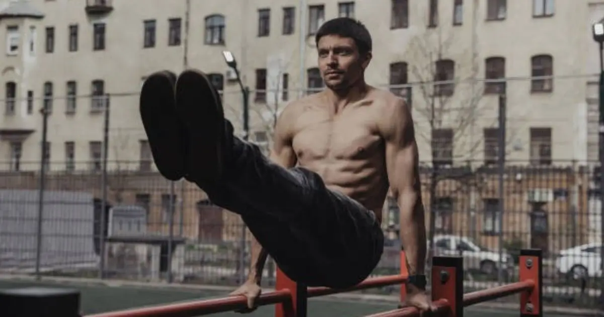 A shirtless man performing a leg raise exercise on parallel bars in an outdoor urban setting.