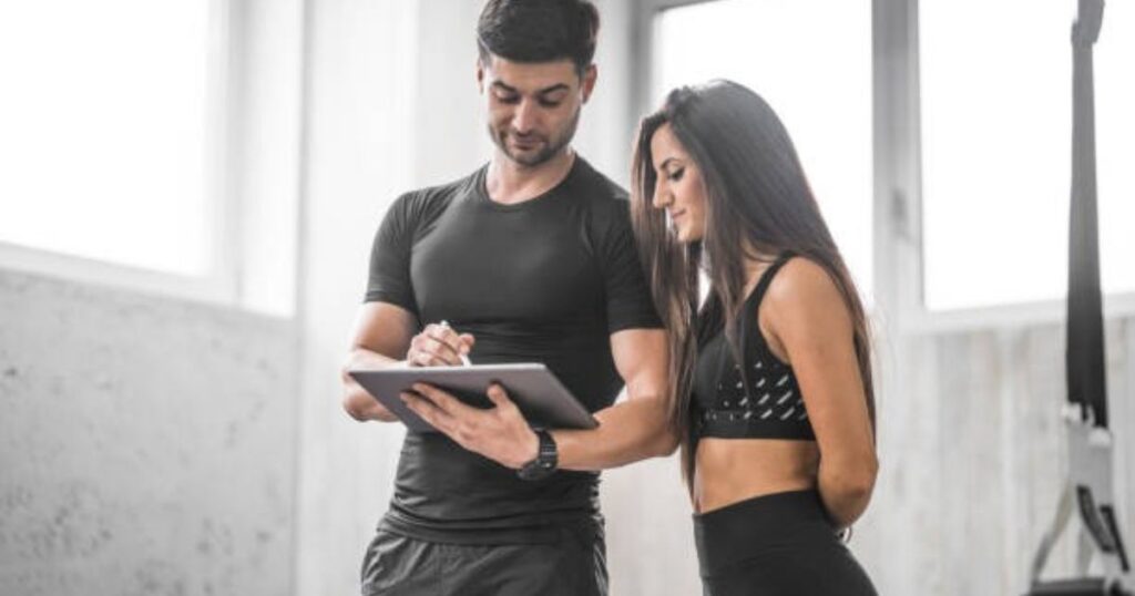 A personal trainer and client are reviewing a fitness plan on a tablet in a bright, modern gym setting.
