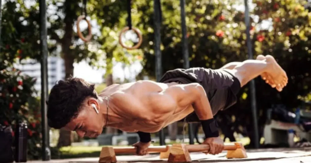 A shirtless man is performing a push-up exercise outdoors on wooden parallel bars, with rings visible in the background.