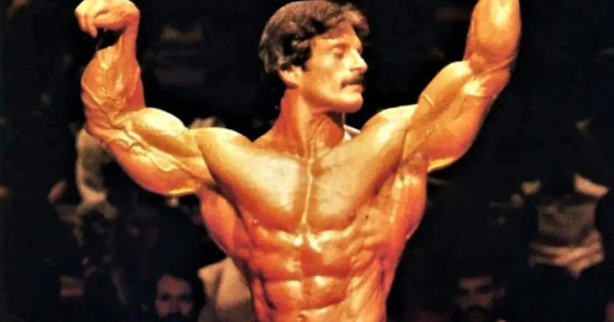 A muscular bodybuilder poses on stage with arms raised, showcasing defined muscles and a powerful physique.
