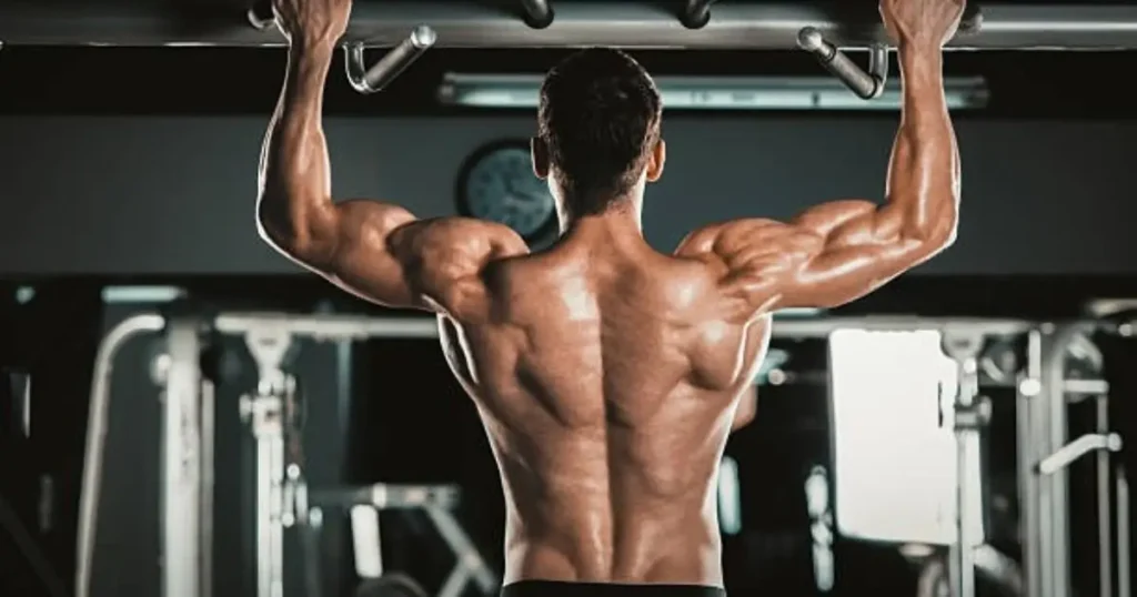 A muscular man is performing a pull-up exercise in a gym, showcasing his defined back and arm muscles. The setting includes exercise equipment and a clock on the wall.